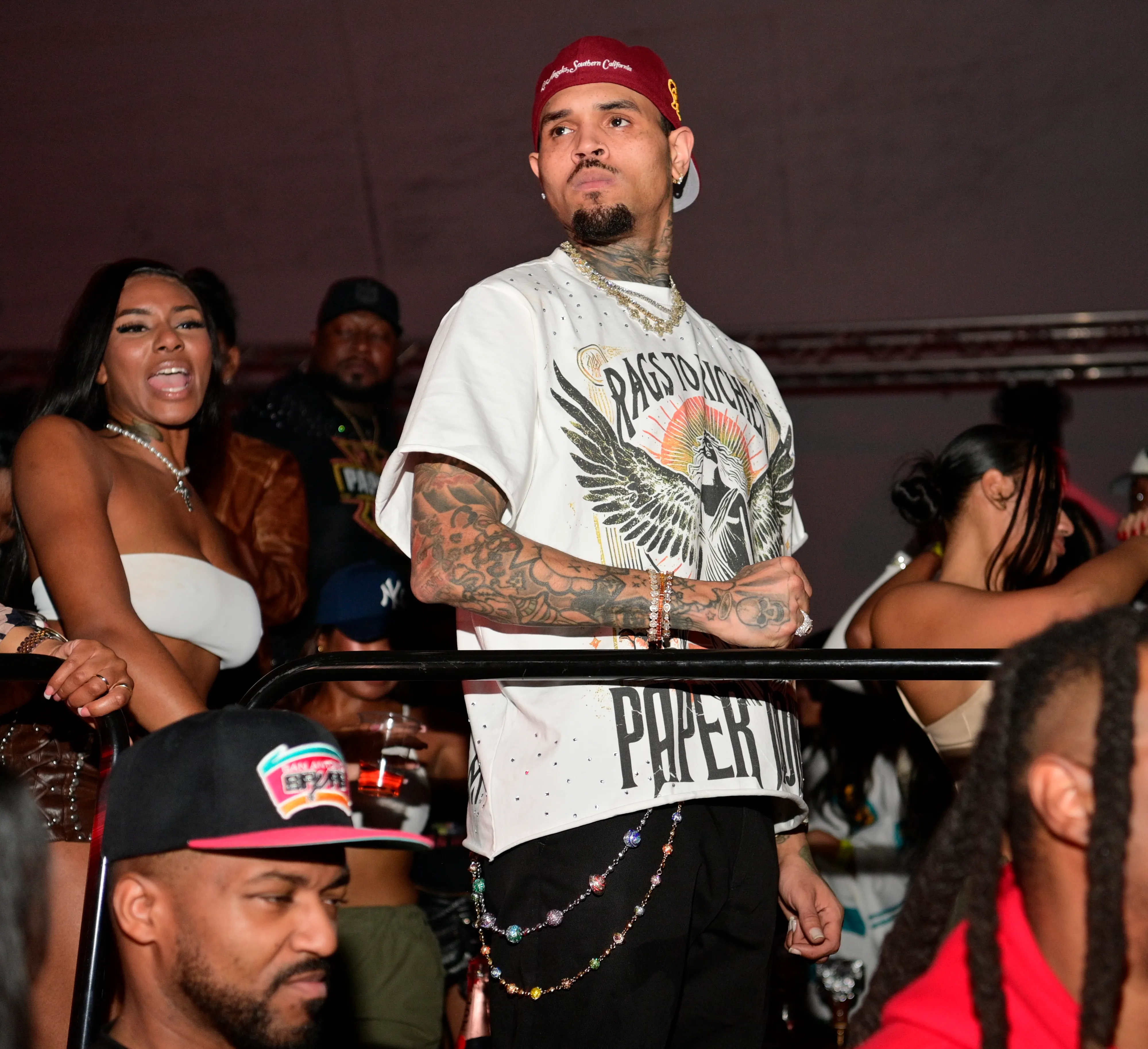 Chris Brown at an event, wearing a white t-shirt and a red cap, with a woman next to him.