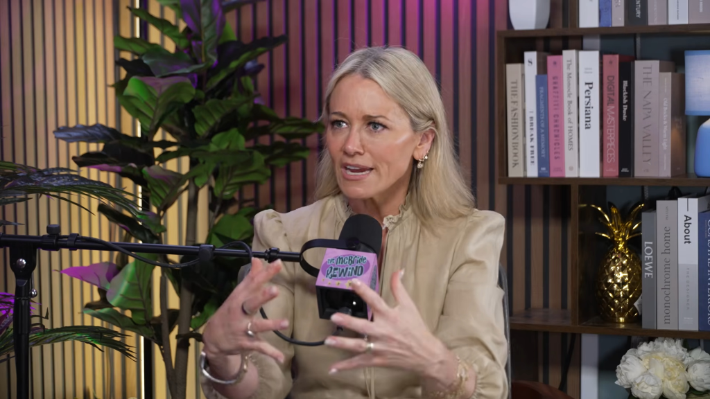 Christine Taylor speaks into a microphone with bookshelves and plants in the background.