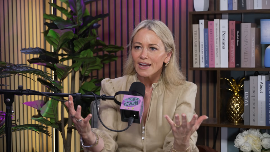 Christine Taylor speaking into a microphone with bookshelves and plants in the background.