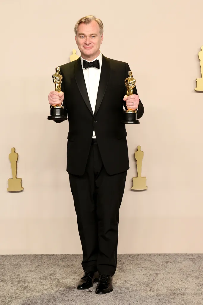Christopher Nolan holding two Oscar awards, wearing a black tuxedo.