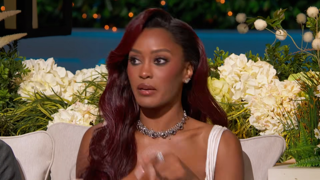 Ciara Miller in a white dress and statement necklace, speaking with a surprised expression.
