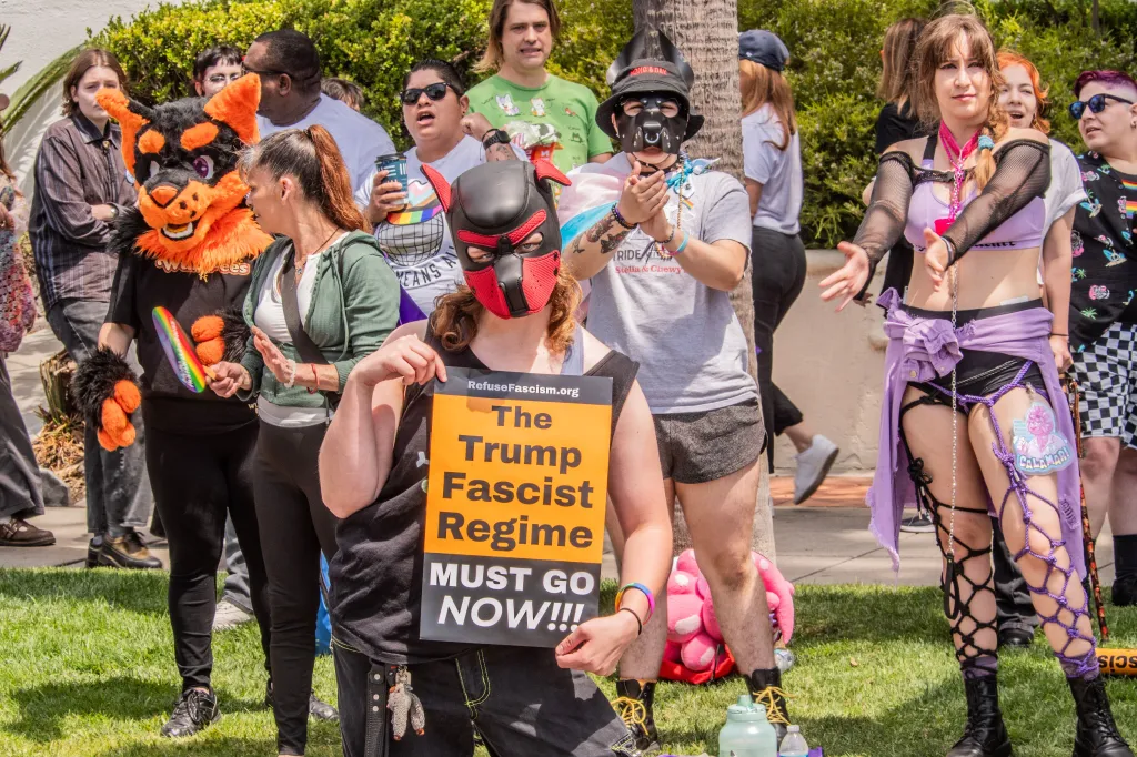 A person in a black and red pup hood holds a sign that reads
