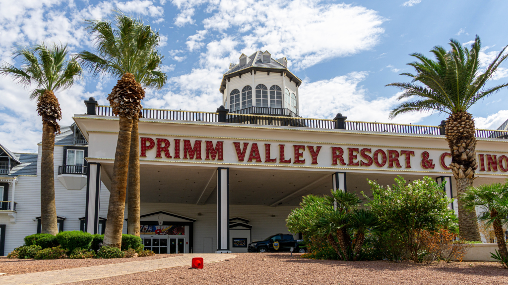 Exterior view of Primm Valley Resort & Casino, a white building with red lettered signage, surrounded by palm trees and desert landscaping.