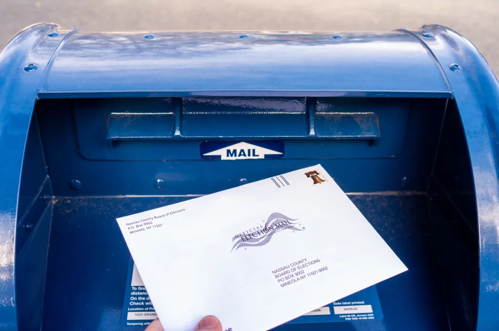 Close-up of a hand placing an absentee election ballot into a blue mailbox in Nassau County, New York.