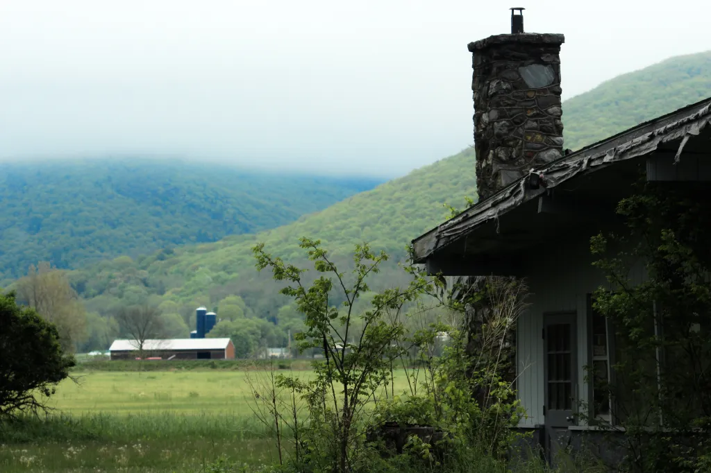 A closed business with a stone chimney and peeling roof in the foreground, with a field, farm buildings, and the Taconic Ridge in the background.