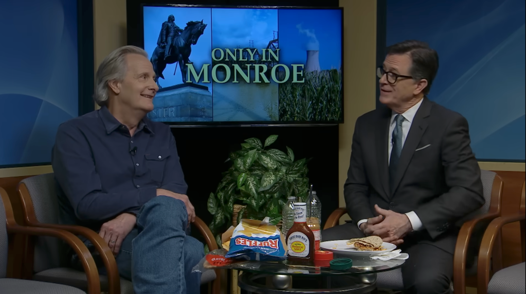Stephen Colbert and a guest sitting on a talk show set with the words 