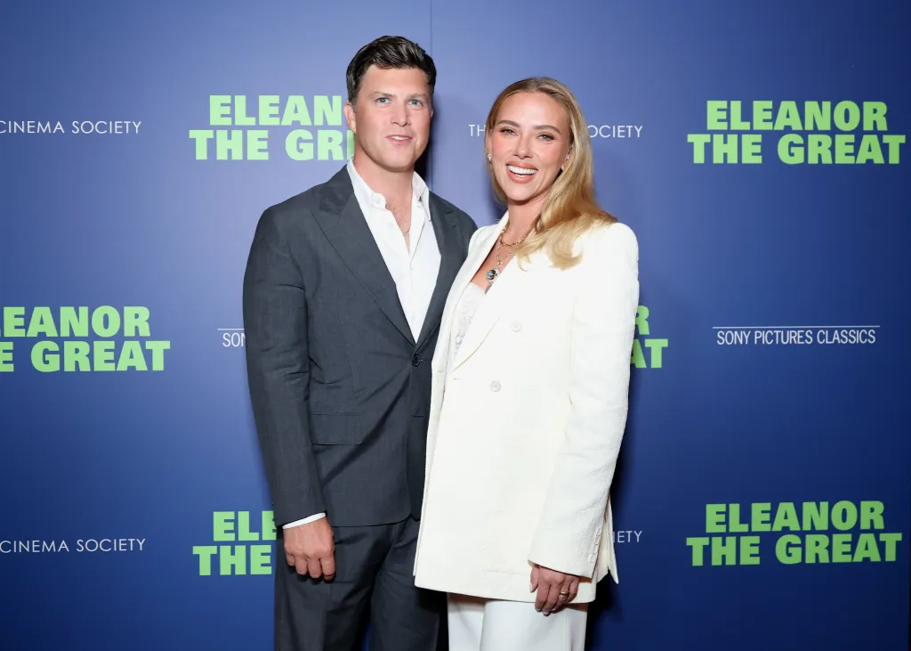 Colin Jost in a dark suit and Scarlett Johansson in a white pantsuit pose in front of a blue backdrop with 