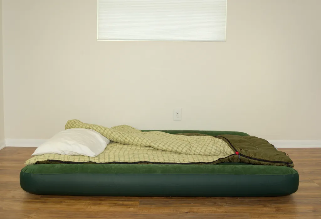 An inflated green air mattress on a wooden floor with a white pillow and an unzipped sleeping bag on top, in a room with light walls and a window with blinds.