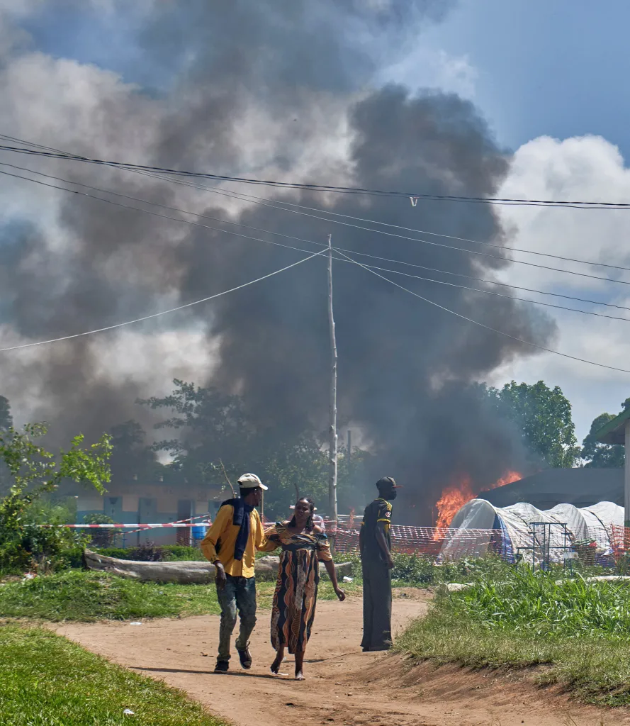 Congolese civilians walk near a burning Ebola treatment center with plumes of smoke rising into the sky.