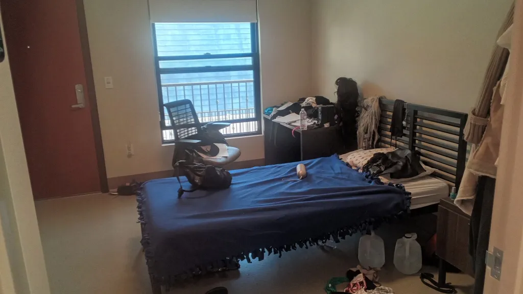 A furnished bedroom with a bed, desk, and office chair, featuring a window overlooking a cityscape.