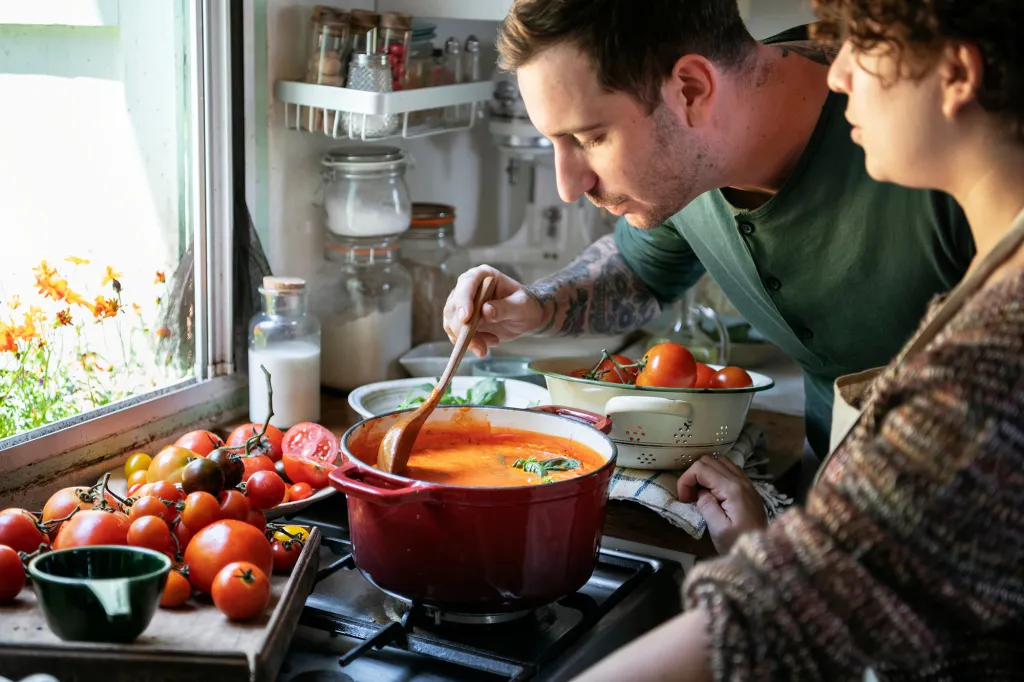 A man stirring tomato soup with a wooden spoon in a pot on a stove next to a woman.