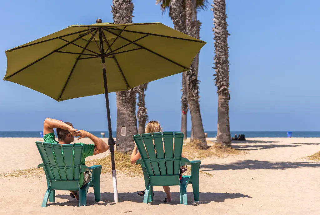 A couple sitting in green chairs under a green umbrella on a sandy beach, looking out at the ocean.