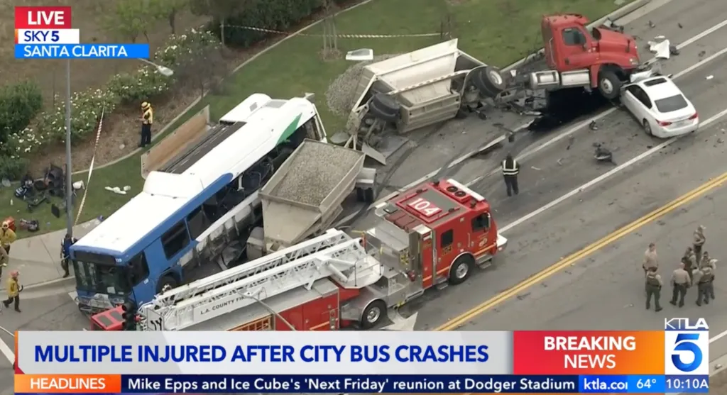 Aerial view of a Santa Clarita Transit bus and an overturned tractor-trailer after a crash, with a fire truck and emergency personnel on the scene.