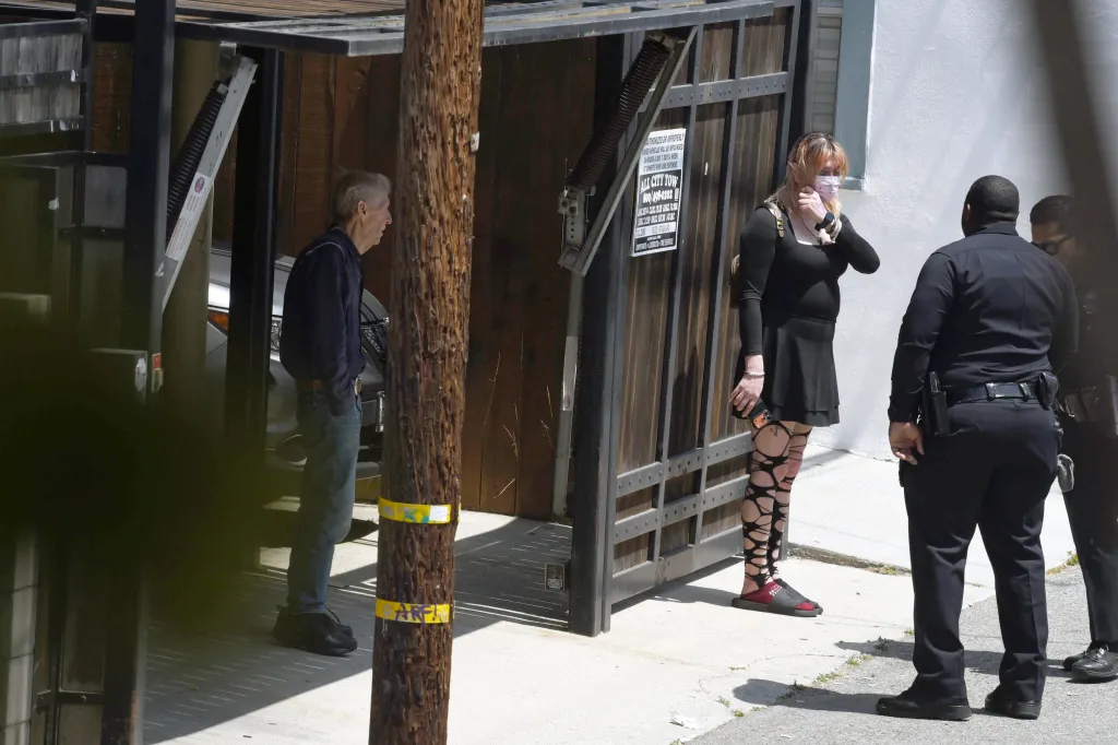 A man, a woman in a mask, and two police officers stand outside a building next to a wooden pole and a sign for 