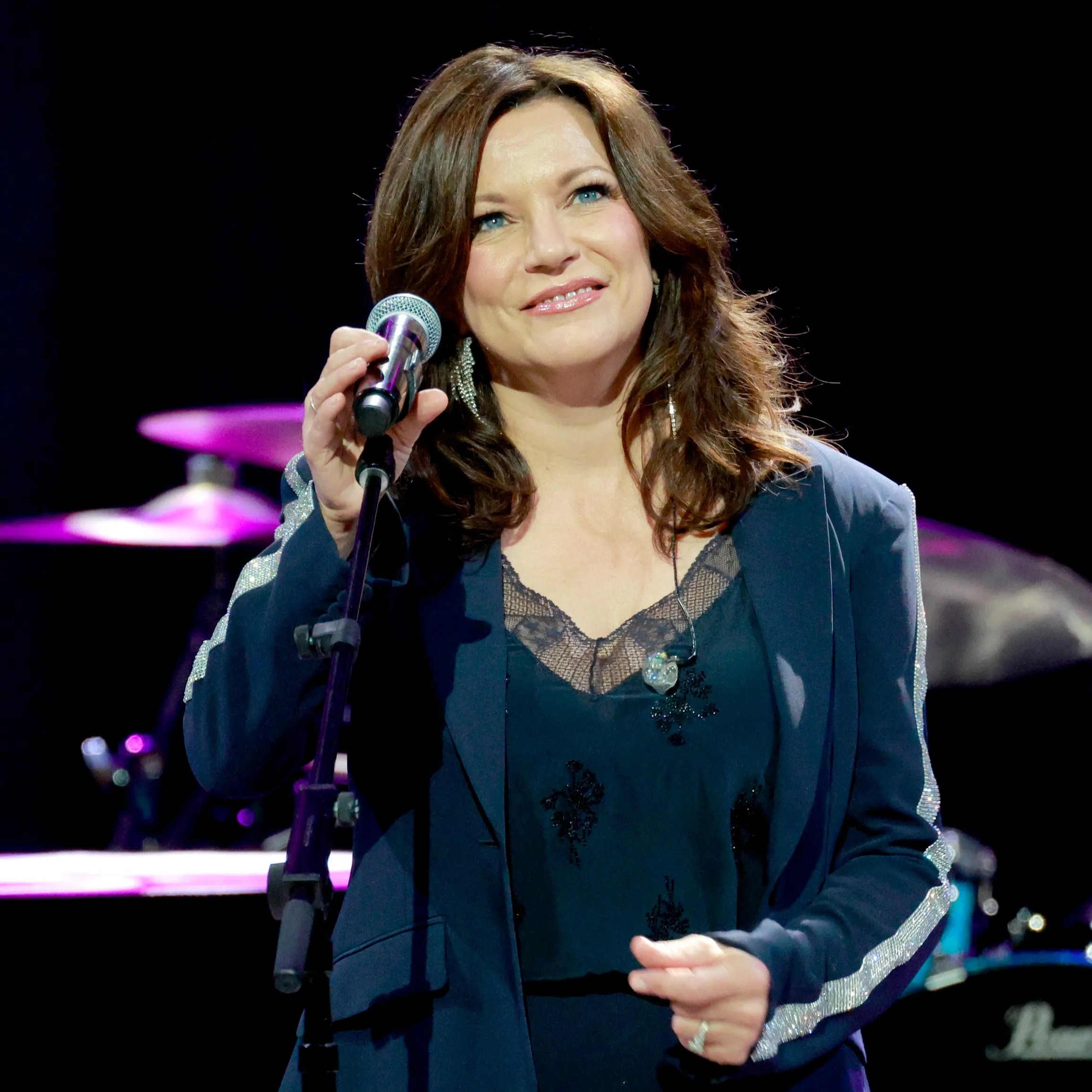 Martina McBride performing on stage, singing into a microphone with a drummer in the background.