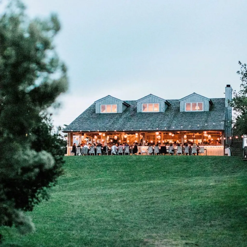 A large building with a covered porch where people are dining, seen from a grassy hill.