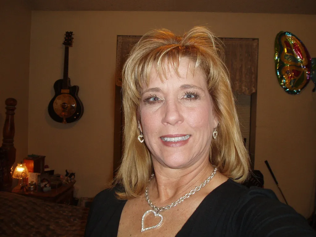 Lynne Alane Shadduck, a blonde woman, smiles at the camera while wearing heart-shaped earrings and a heart-shaped necklace.