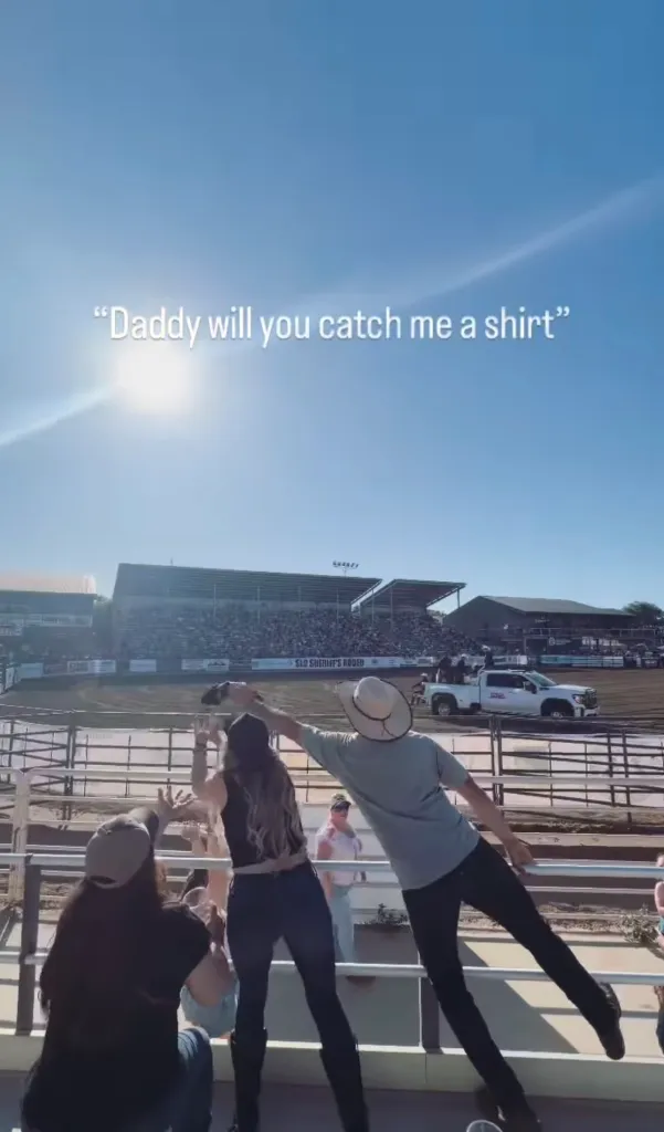 A man in a cowboy hat catches a T-shirt thrown from the rodeo arena while a woman next to him reaches for it, under a bright blue sky.