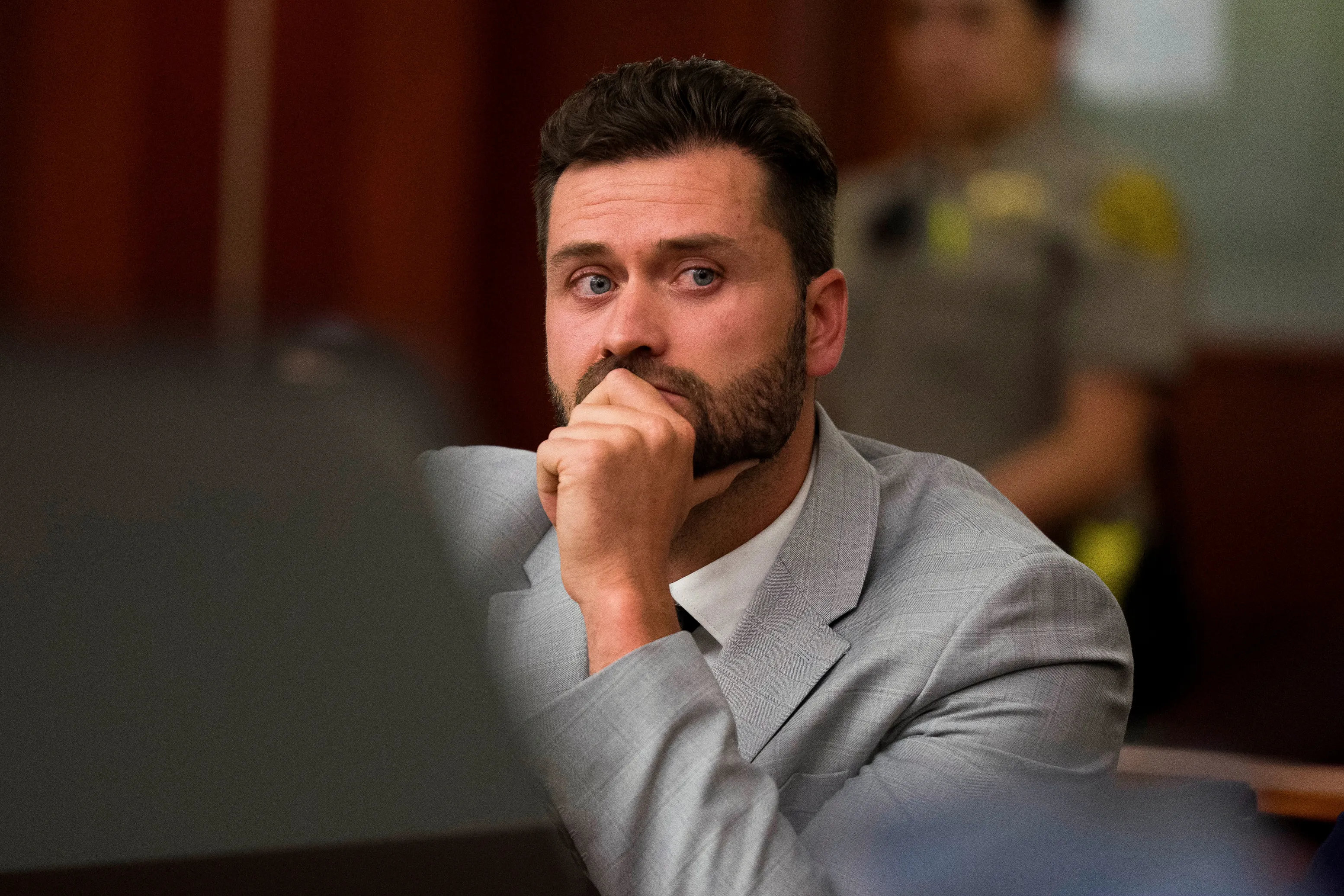 Dakota Mortensen in a courtroom, wearing a gray suit jacket and white shirt, with his hand on his chin.