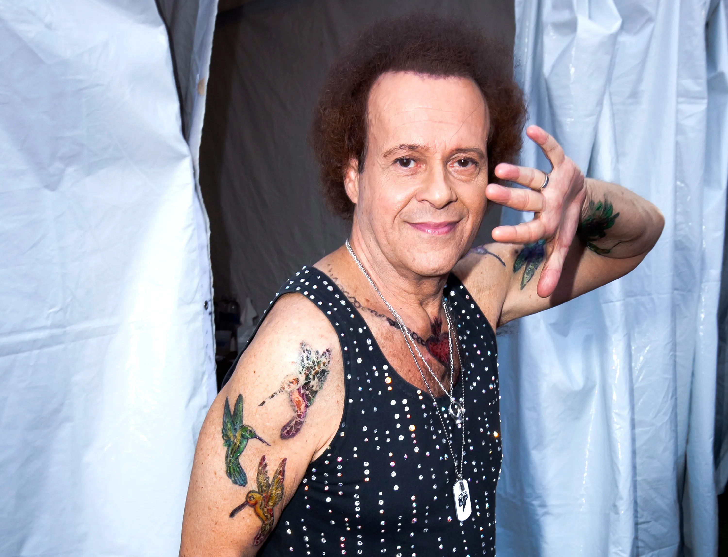 Richard Simmons poses in a black sequined tank top, showing off his tattoos, and smiling at the camera.