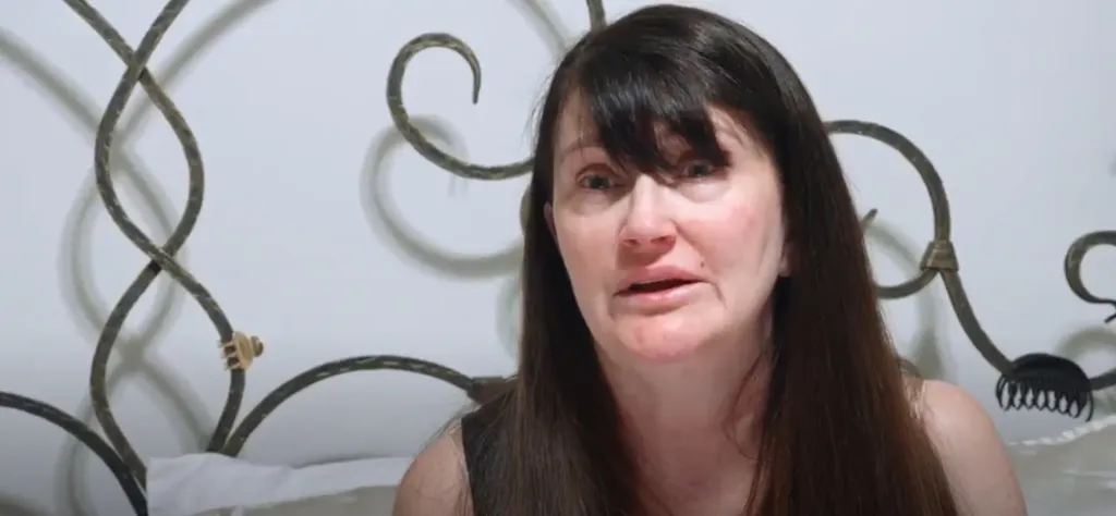 Debby, a woman with long dark hair, appears worried or upset while sitting in front of a white headboard with decorative metalwork.