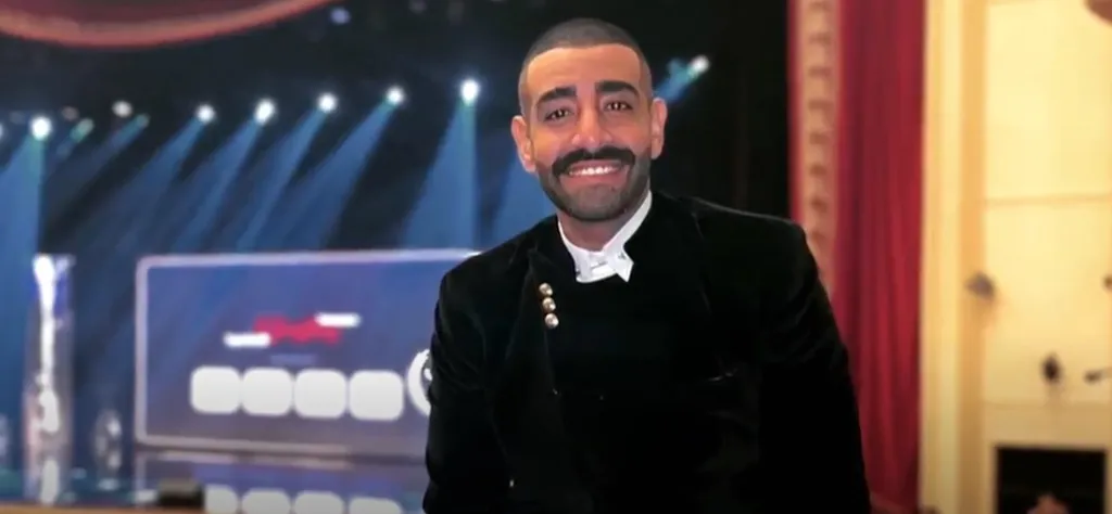 A man with a mustache and beard in a black suit smiles against a stage background.