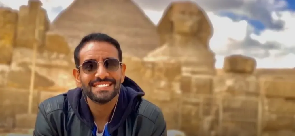 A man with sunglasses and a beard smiling in front of the Sphinx and a pyramid.