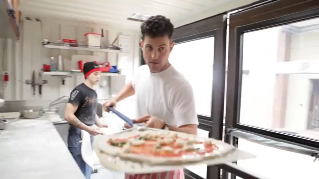 A man in a white shirt and red striped apron holding a raw pizza on a peel, with another man in a black shirt and beanie in the background.