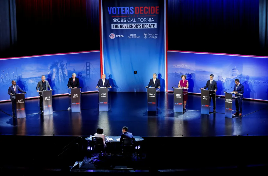 Eight candidates for California governor stand at podiums on a debate stage, with two people at a table in the foreground.