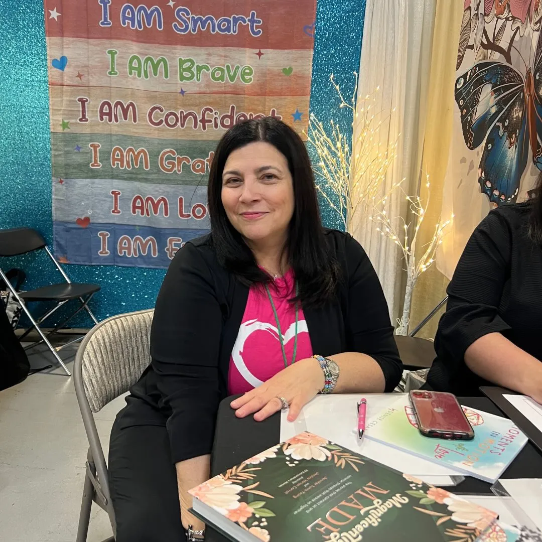 Denise Cesare smiling while sitting at a table with books, with a banner behind her that reads 