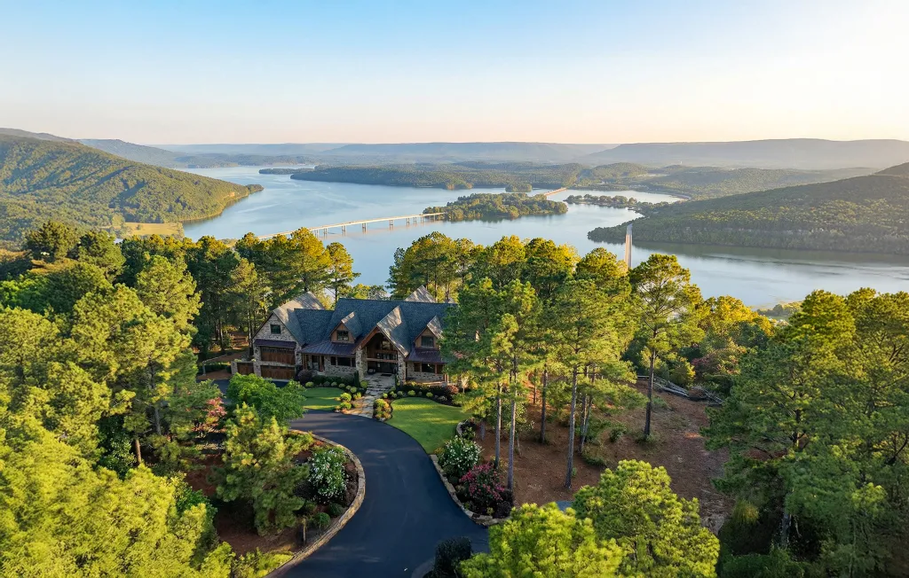 A stone house with a long driveway surrounded by green trees, with a large lake and mountains in the background.