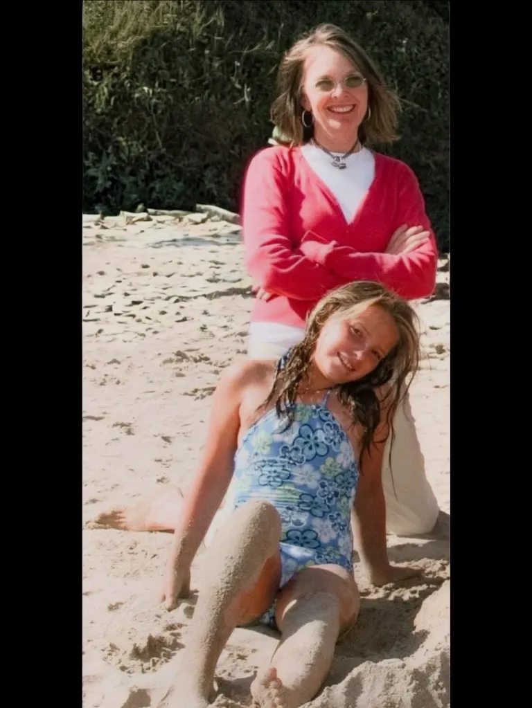 Diane Keaton smiling with her arms crossed behind her daughter, Dexie White, who is sitting on the sand in a swimsuit.