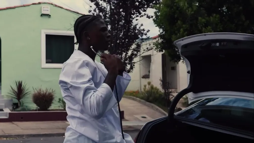 Dominic Antoine Jr. wearing a white coat and holding a stethoscope, standing beside an open car trunk.