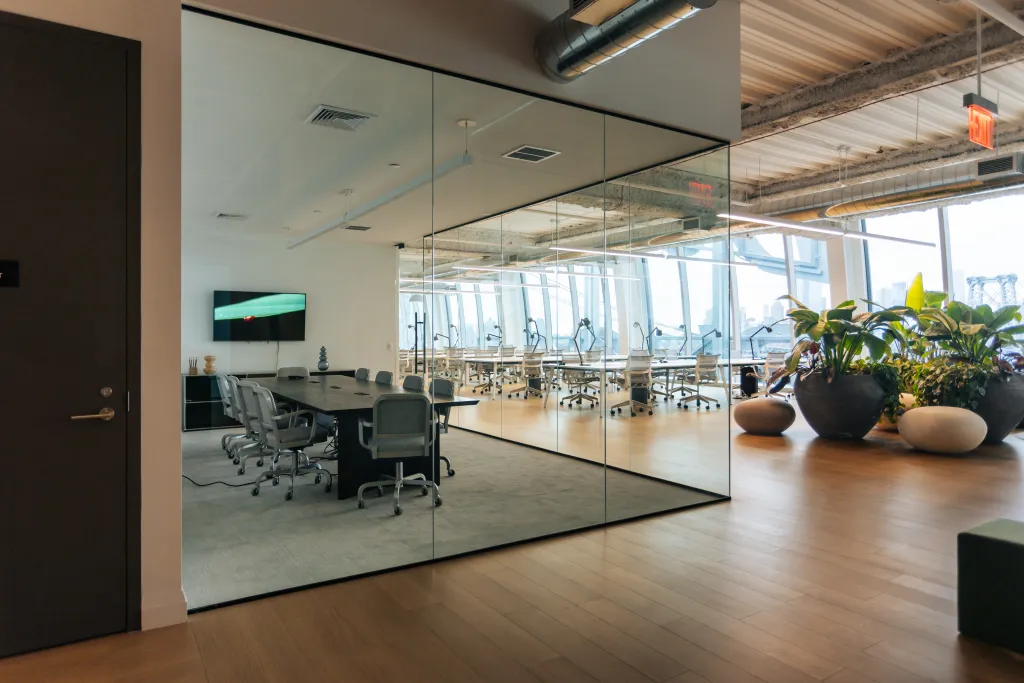 A conference room with a long table and chairs separated by a glass wall from a large office space with floor-to-ceiling windows and plants.