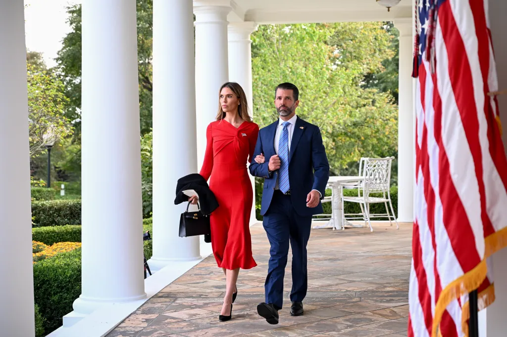 Donald Trump Jr., in a blue suit, and Bettina Anderson, in a red dress, walk past an American flag.