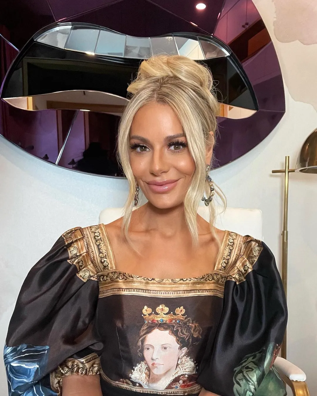 Dorit Kemsley smiling and posing in a black and gold dress with puffy sleeves and a portrait of a woman wearing a crown.