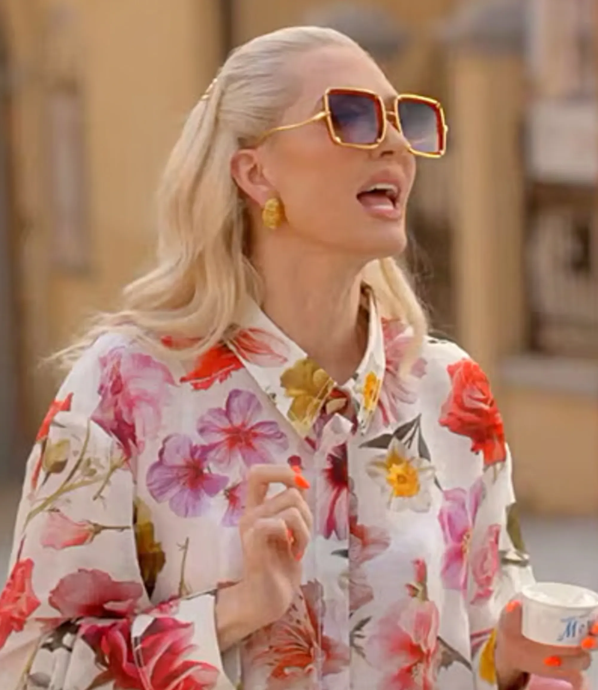 Erika Jayne wearing a floral shirt and sunglasses, holding a small cup.