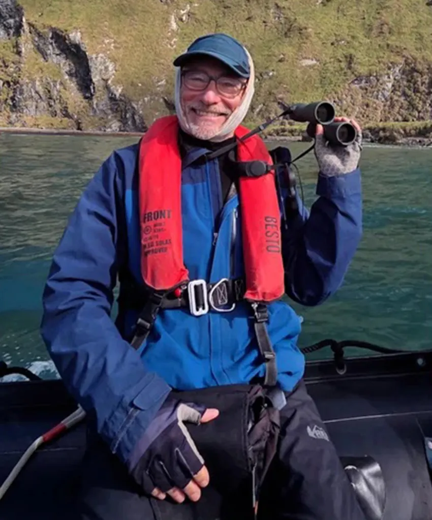 Dr. Stephen Kornfeld, wearing a life vest and a cap, with bandages around his face, holds binoculars while on a boat.
