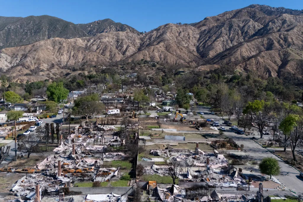 Aerial view of a residential area in Altadena, California, showing workers cleaning debris from homes destroyed by the Eaton Fire, with dry, barren mountains in the background.