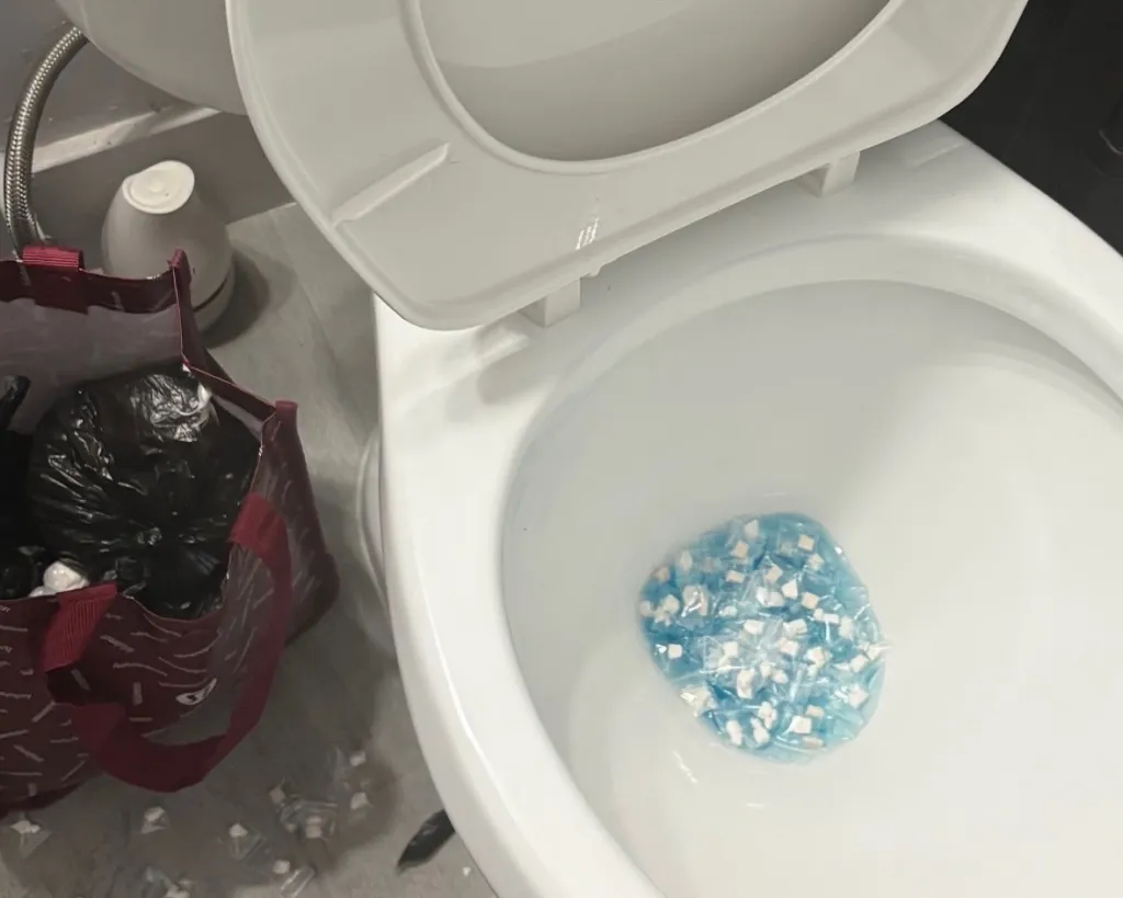 A toilet with blue-wrapped drug packets in the bowl and a bag of similar packets on the floor next to it.