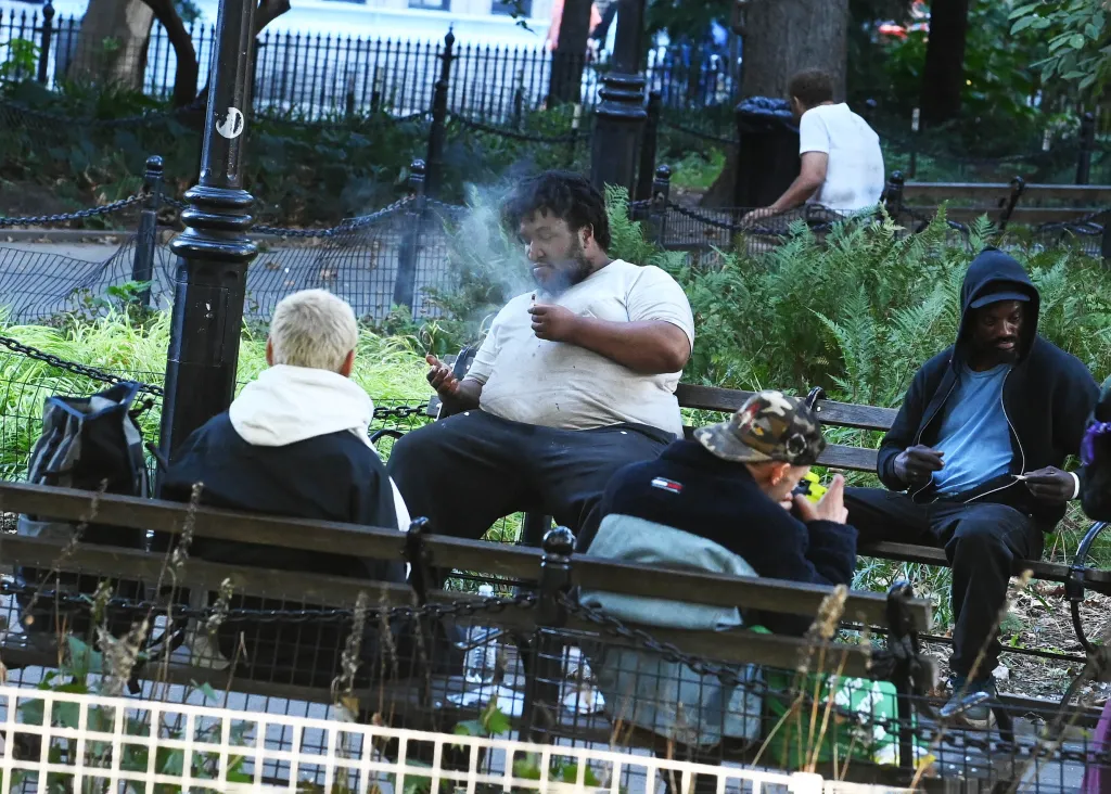 People sitting on a park bench, one smoking a pipe, surrounded by greenery.