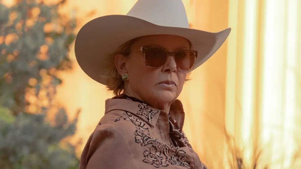 An older woman wearing a white cowboy hat, sunglasses, and an embroidered western shirt looks off-camera in warm golden light outdoors.