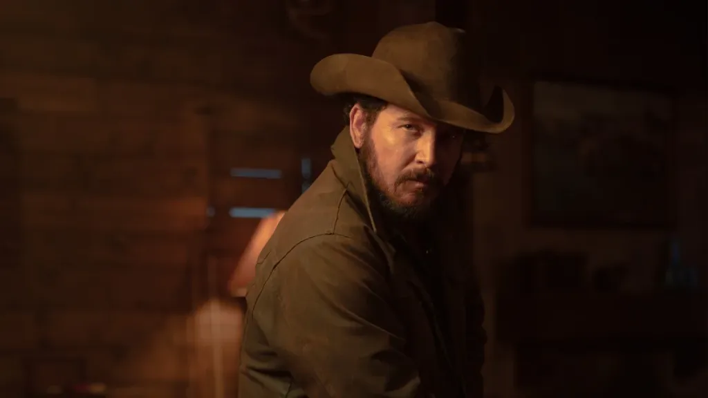 A rugged man in a cowboy hat and dark jacket stands in a dimly lit rustic cabin, looking intensely toward the camera under warm amber lighting.