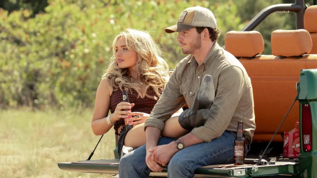 A man and woman sit side by side on the open tailgate of a vintage green Ford Bronco in a dry, grassy field. The woman, with long blonde hair, holds a drink, while the man wears a tan baseball cap and a gray button-down shirt. Beer bottles rest beside them. The scene evokes a relaxed, rural Texas setting. 