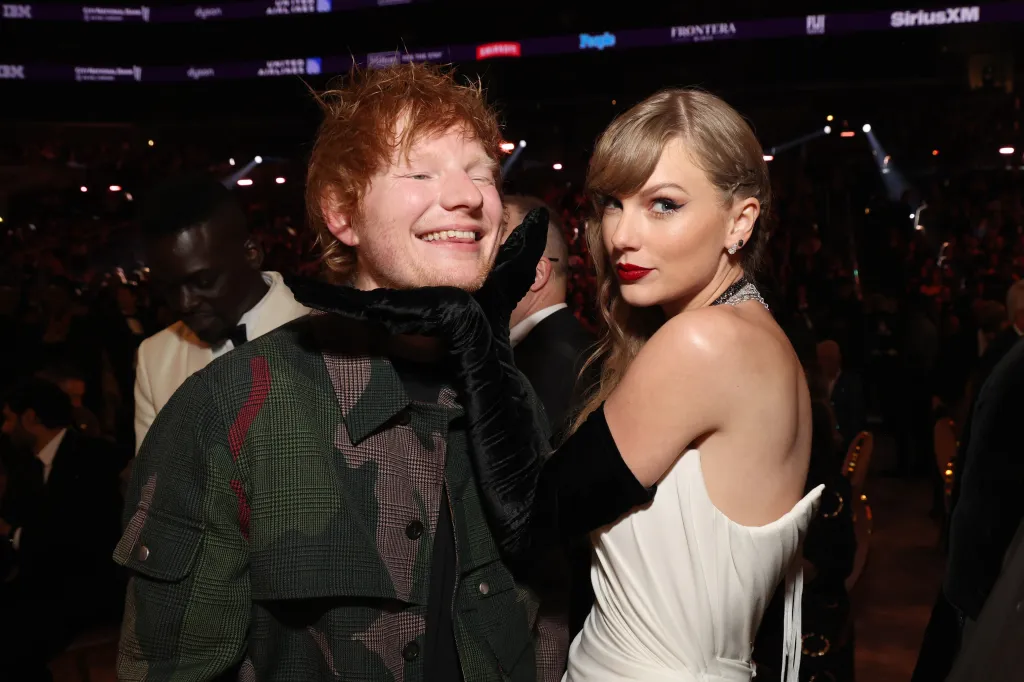 Taylor Swift in a white dress playfully holding Ed Sheeran's chin, who is smiling broadly, wearing a camouflage jacket, at the 66th GRAMMY Awards.