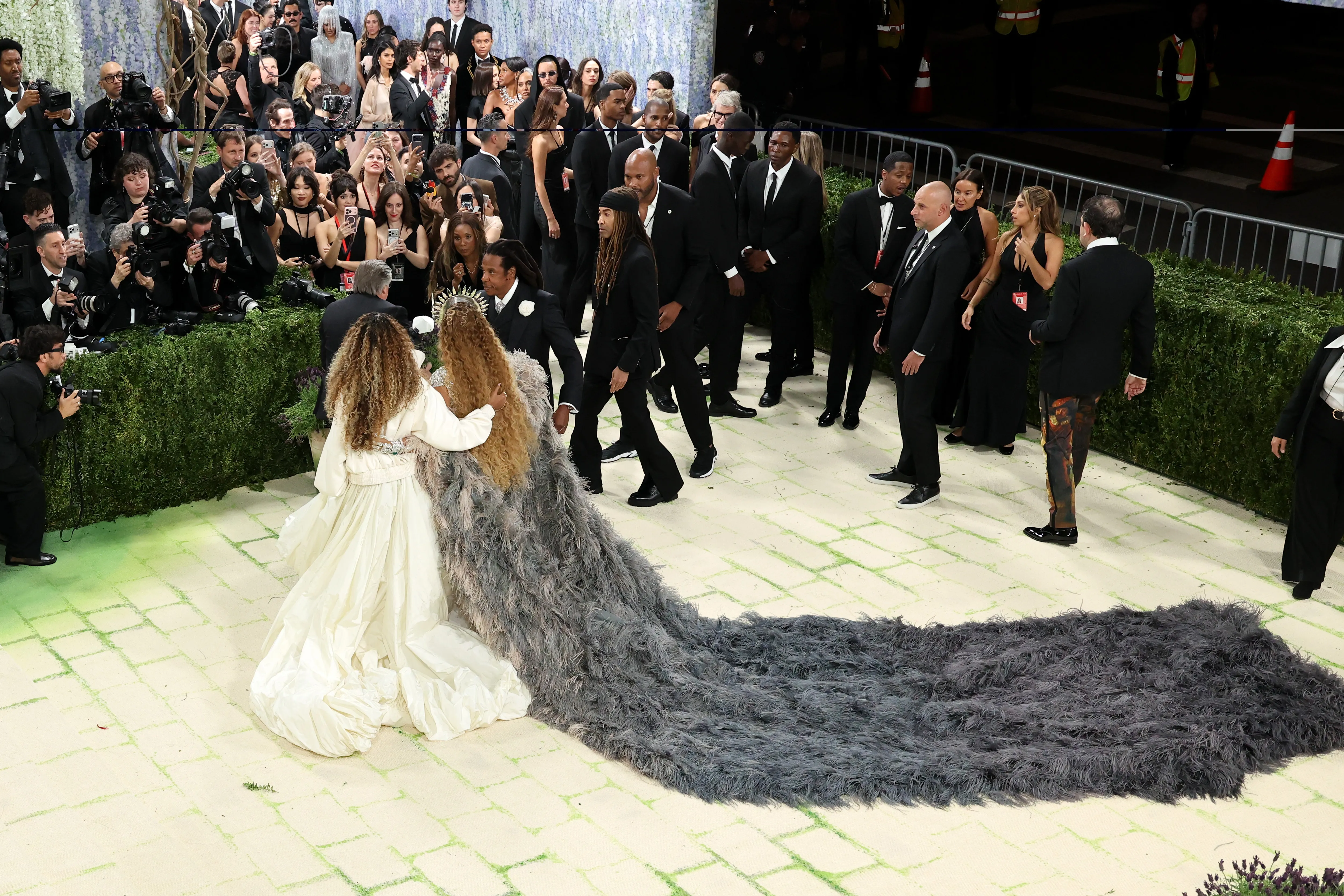 Blue Ivy Carter and Beyoncé walk the Met Gala red carpet, with Beyoncé wearing a dramatic gray feathered train, surrounded by photographers and attendees.
