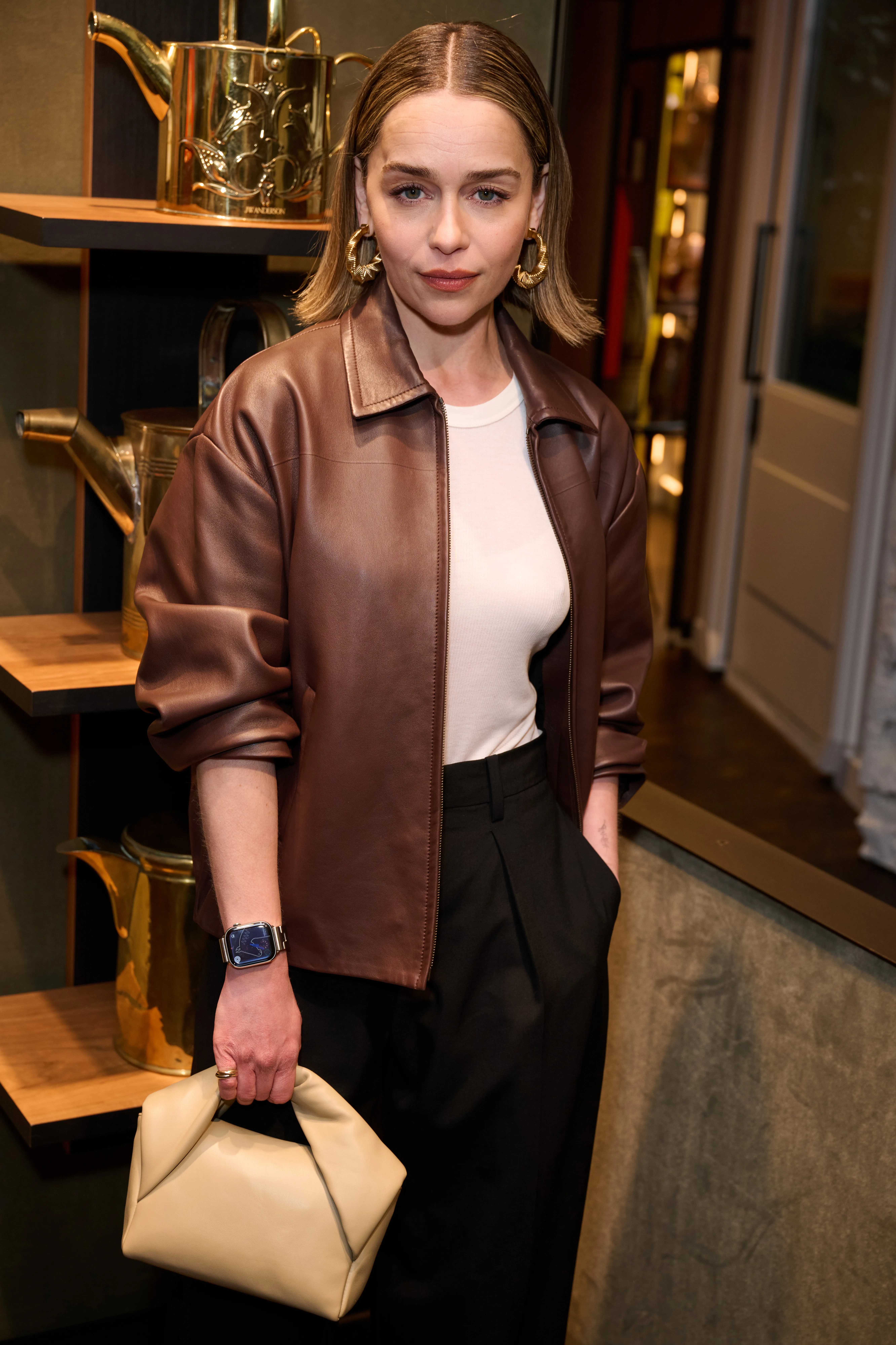 Emilia Clarke wearing a brown leather jacket, a white shirt, black pants, gold earrings, a watch, and holding a small cream-colored handbag.