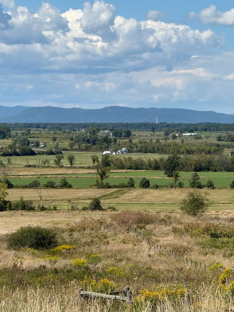 The Fort Edward Solar footprint area shows a vast expanse of green and brown fields with scattered trees and buildings, bordered by distant mountains under a cloudy sky.