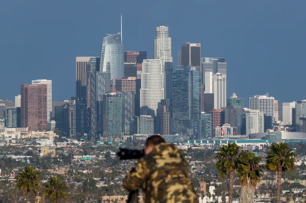 A person taking a photograph of the Los Angeles downtown skyline, which features prominent buildings like the U.S. Bank Tower, Wells Fargo, AON, and Deloitte.
