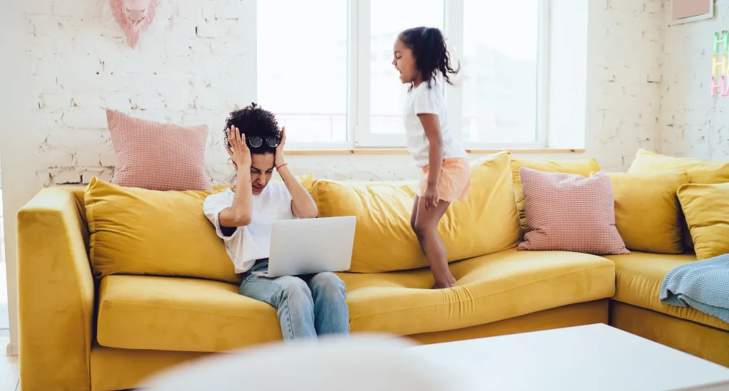 An annoyed mother holds her head while trying to work on a laptop as her daughter jumps and screams on the couch.